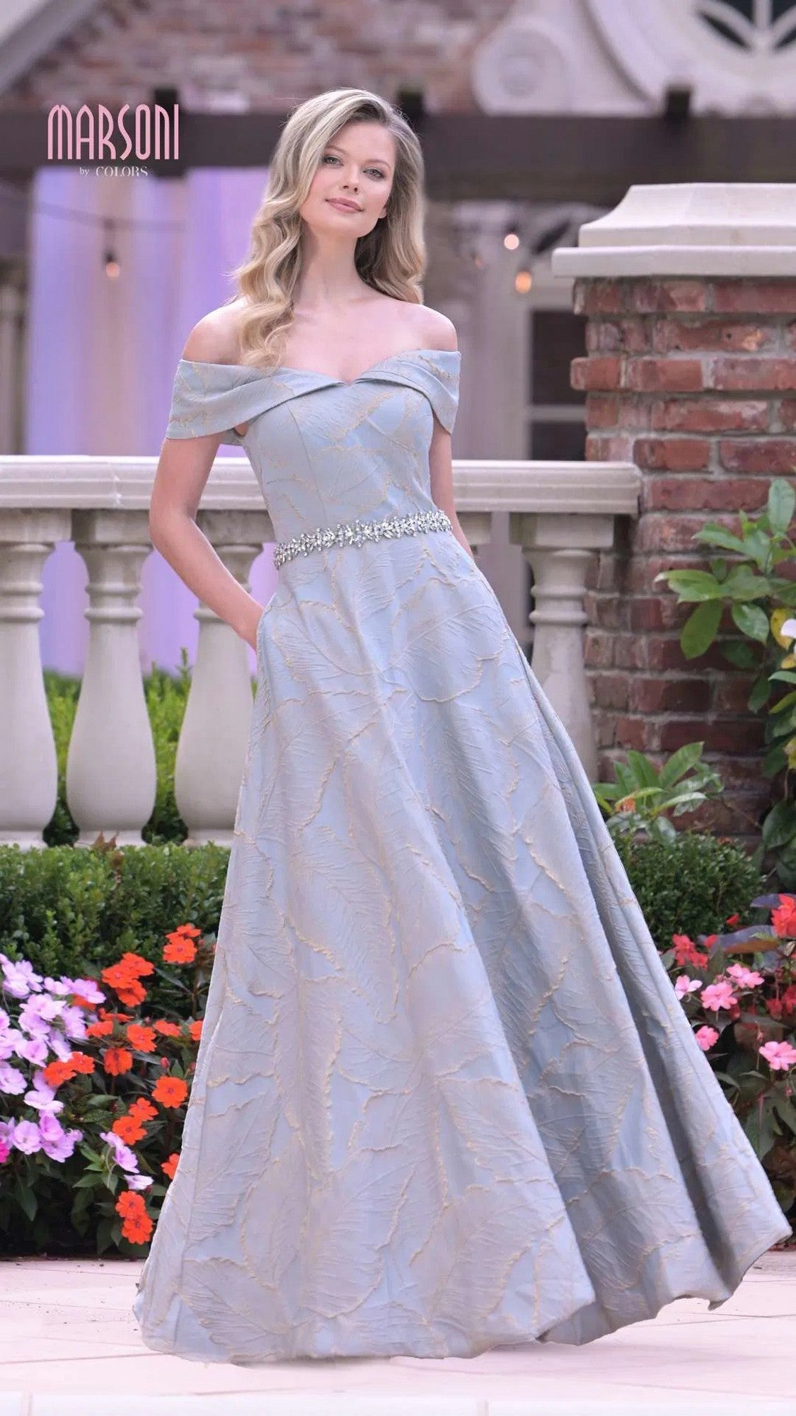 A woman stands outdoors near flowers and brick columns, wearing the Marsoni MV1265 long formal dress in Seaglass—a pale blue, off-the-shoulder A-line gown with a beaded waistline flowing gracefully to the floor.
